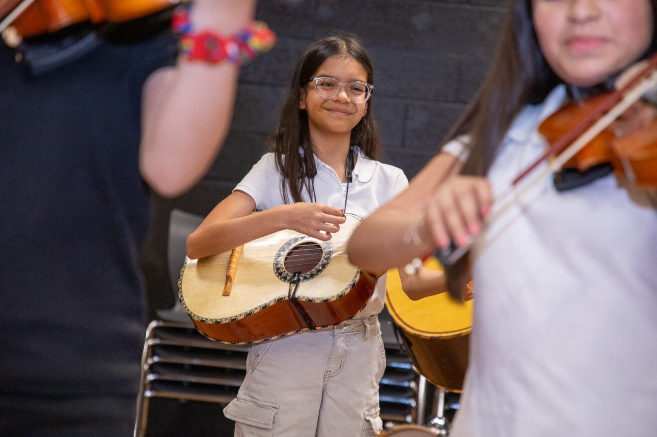 A little girl with long brown hair and glasses smiles with her guitar