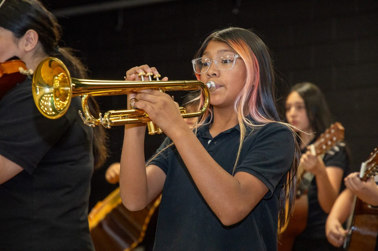 A girl with long black hair and glasses plays her trumpet