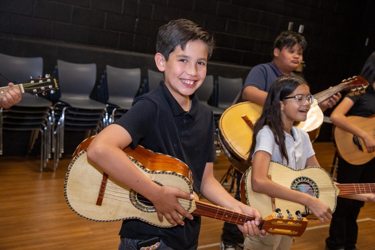 A little boy with dark brown hair smiles big with his guitar